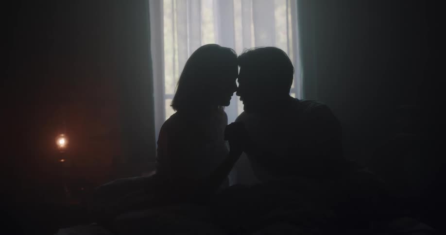 Silhouette of a young couple touching foreheads on the bed inside a dimly lit room