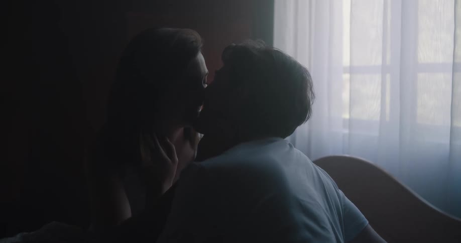 A man and a woman in a room kissing with a wall and window in the background