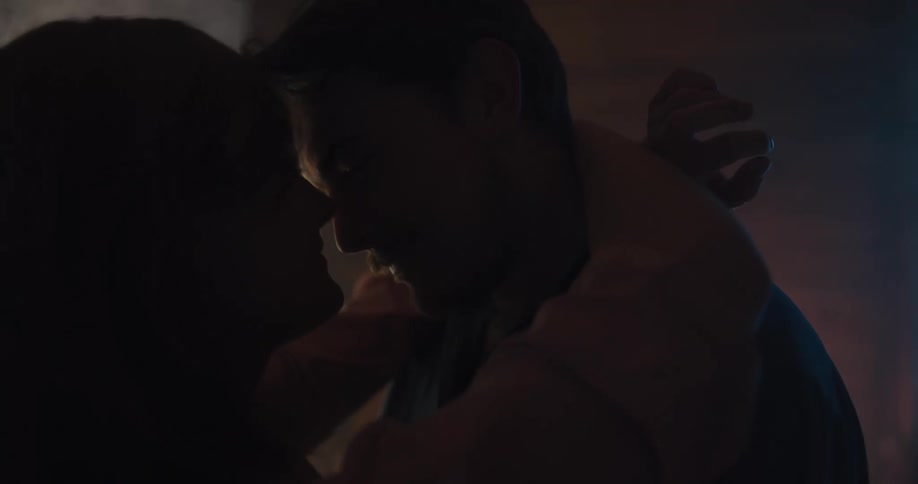 A man and woman embrace and kiss while standing before a fireplace in a dimly lit room