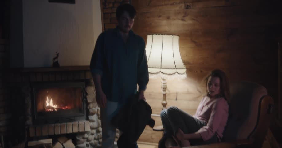 A man and a woman in a room with the woman sitting on a chair with a lamp and a fireplace in the background