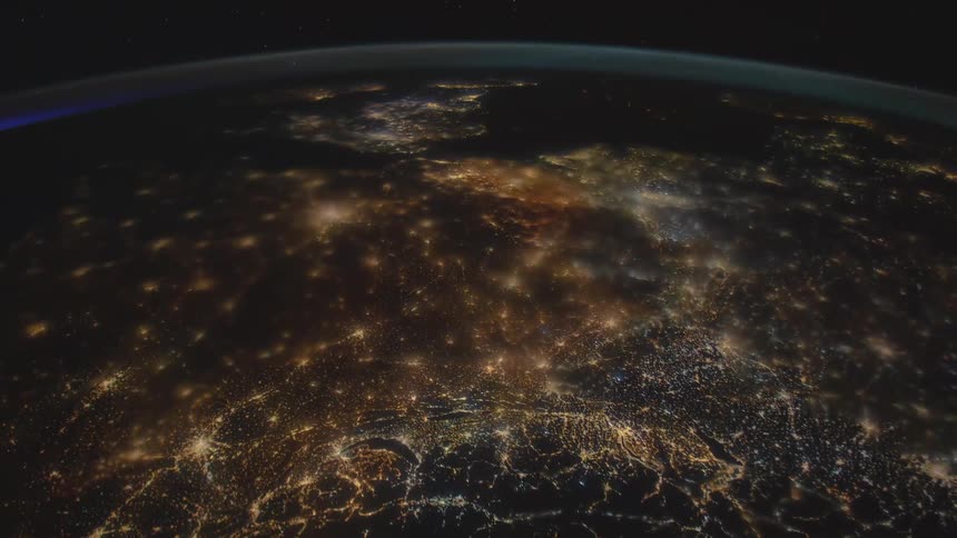 The surface of the earth can be seen below and areas populated by people are illuminated in the darkness