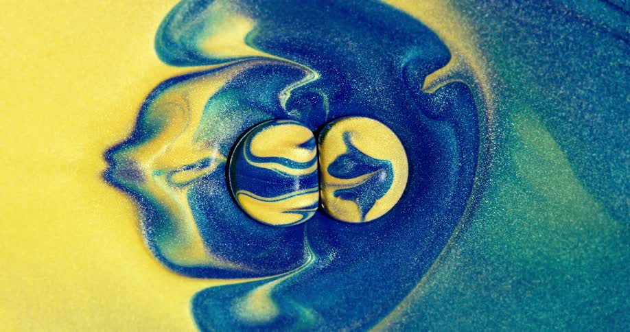 A mixture of yellow and blue liquids intermingle as multicolored droplets bump into each other on the surface