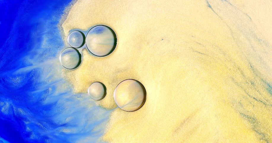 A liquid with blue and yellow coloration and many glittery particulates in it flows and is mixed around some large droplets on its surface