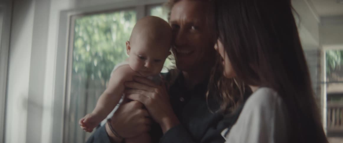 A man and a woman in a room with the man holding a baby with a door and window in the background