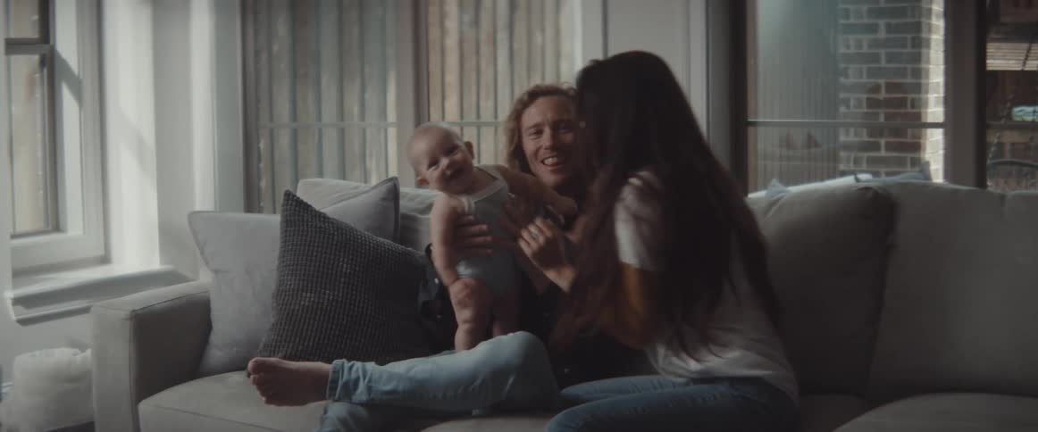 A woman and man sit on a couch playing with an infant and laughing together together