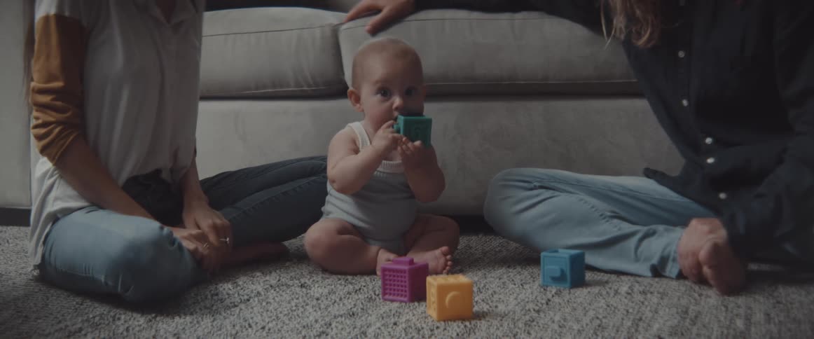 A baby chews on a plastic block while sitting on a rug with his parents at home