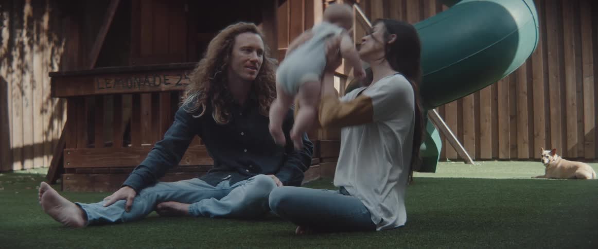A woman and a man sitting on the ground with the woman playing with a baby with a fence and a dog in the background
