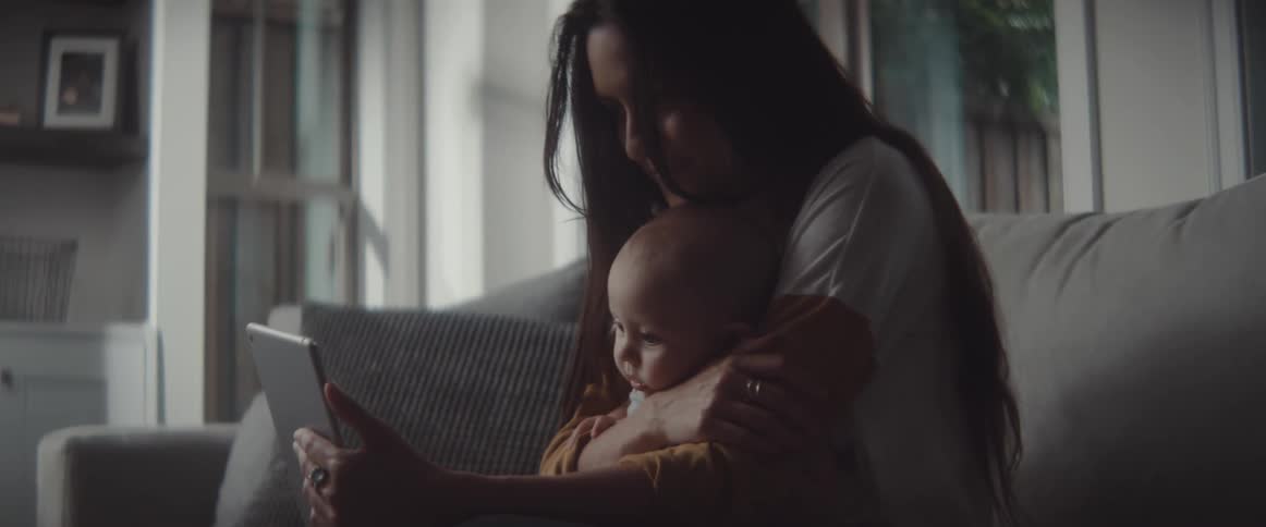 A woman sitting on a sofa in a house kisses the head of a baby being held in her lap as she and the baby look at the screen of a tablet