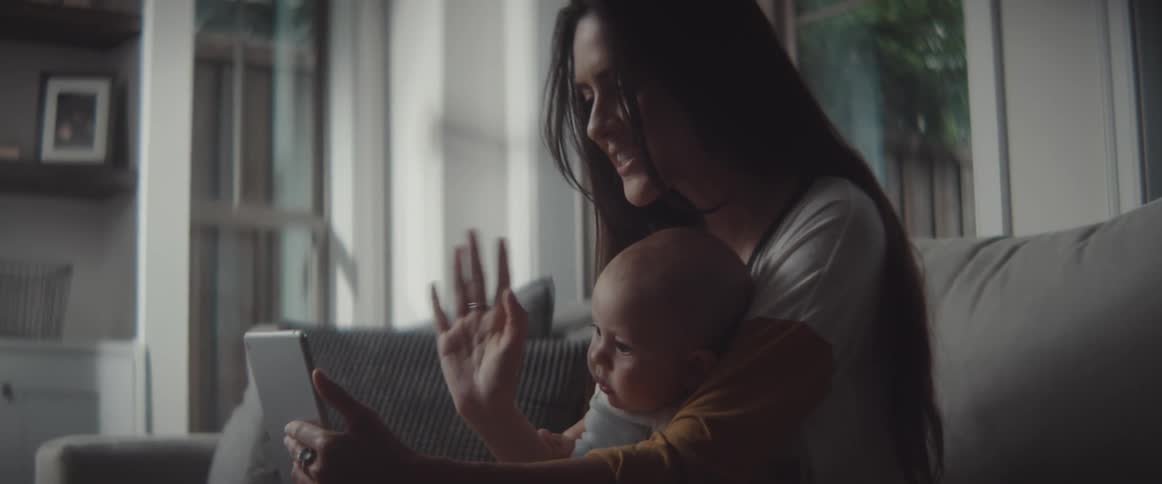 Woman with long dark hair and sweatshirt holds baby and makes a video call on tablet in the living room at home