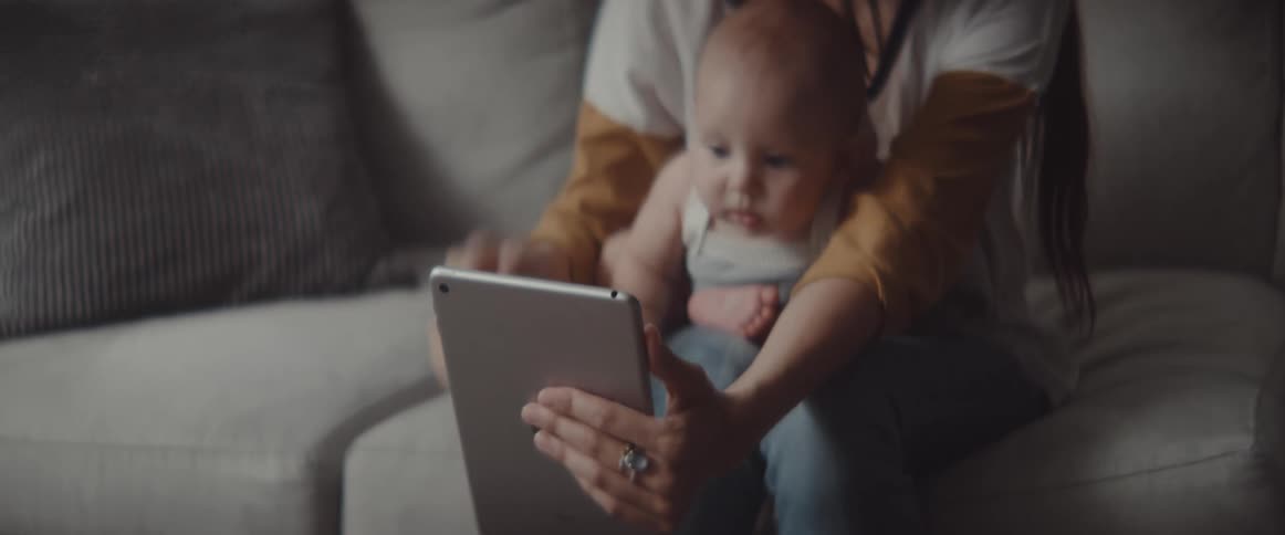 Mother holds child in lap while video calling on tablet and helps child wave at screen