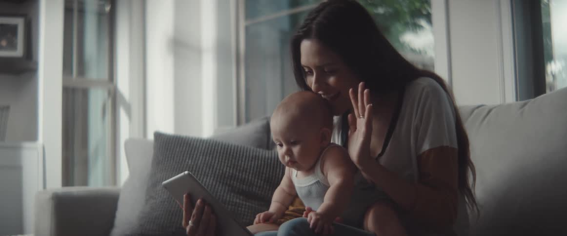 A woman in a room sitting on a couch holding a baby and a tablet
