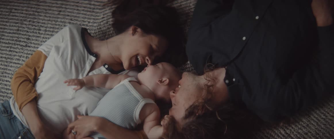 Mother and father lie on backs with baby between them smiling and laughing as baby touches each of them in turn