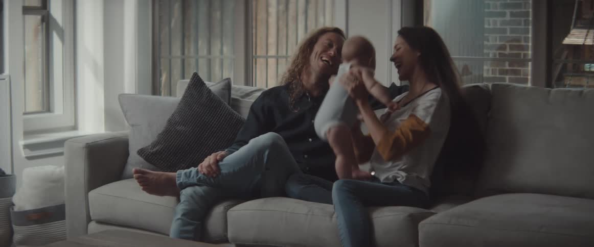A man and woman sit on a couch and laugh as a woman lifts her infant up in the air