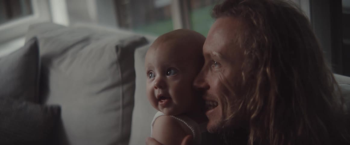 Father and baby smile as father points while they both occupy couch