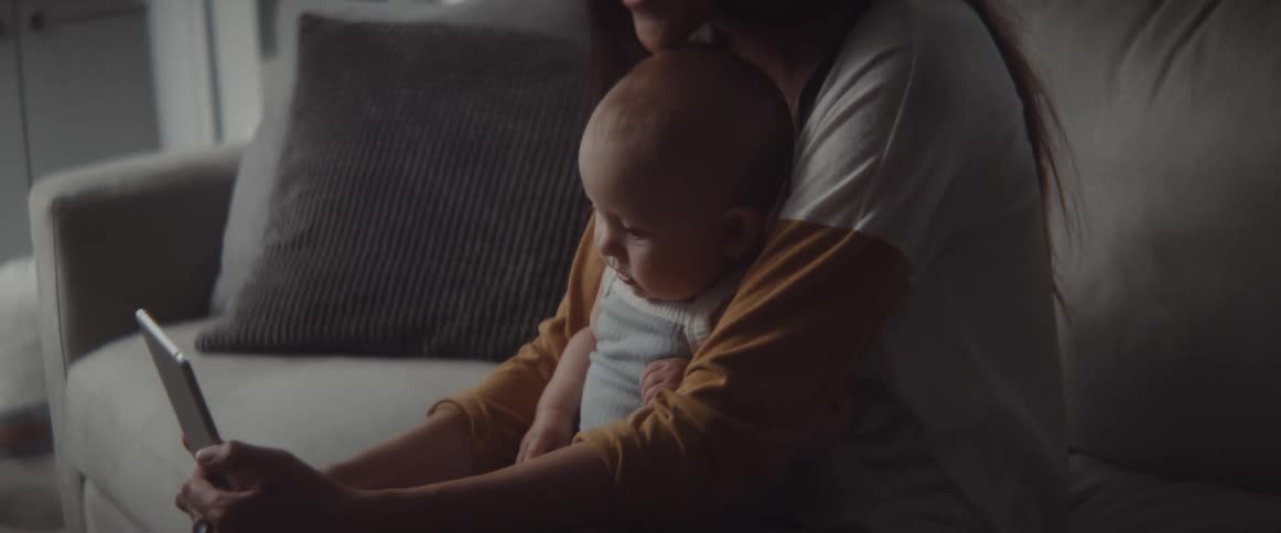 A woman sits on a couch with a baby and has a video call on a tablet