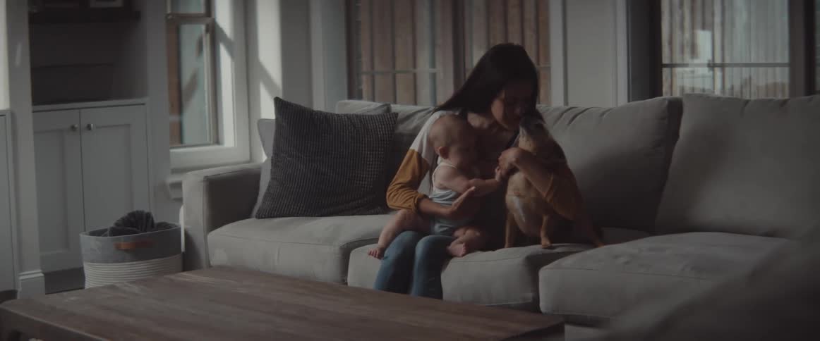 A woman sits on the couch with her infant son while teaching him how to pet the family dog