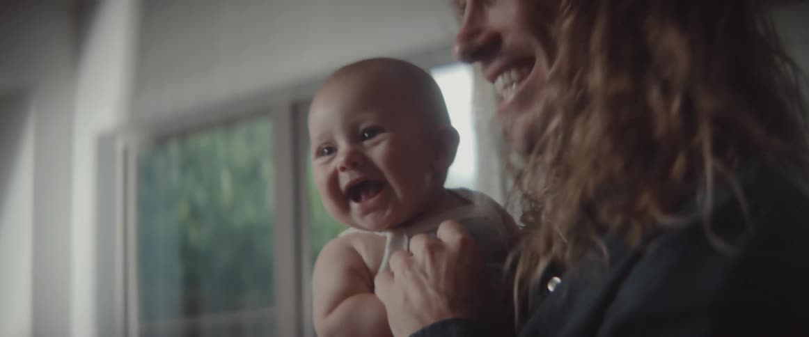 Father with long blonde wavy hair holds baby son in the living room at home
