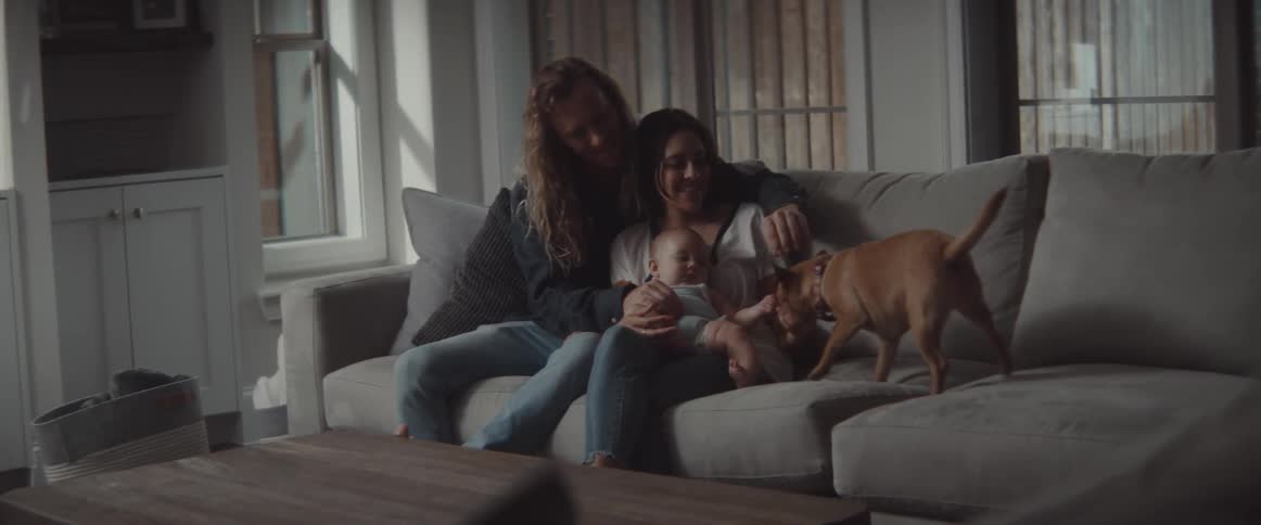 Father with wavy long hair and brunette mother hold baby and caress small dog in the living room