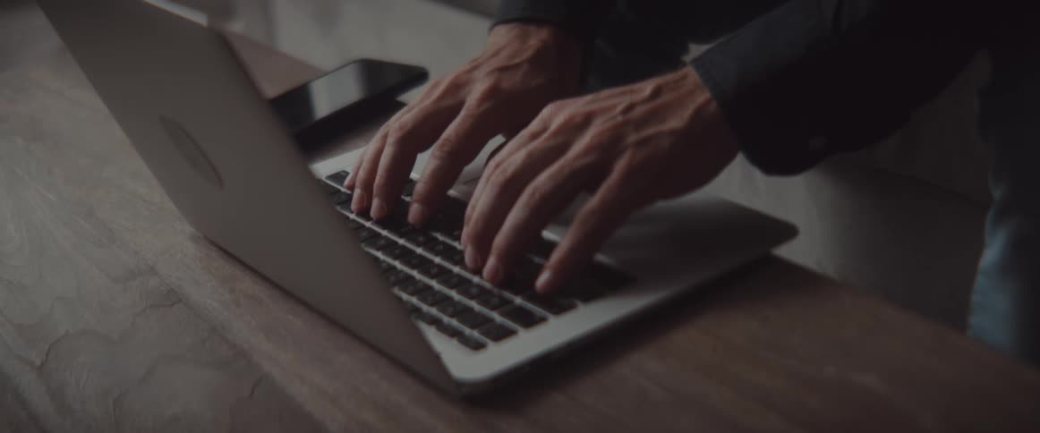 A person stands at a table leaning over to type on a laptop
