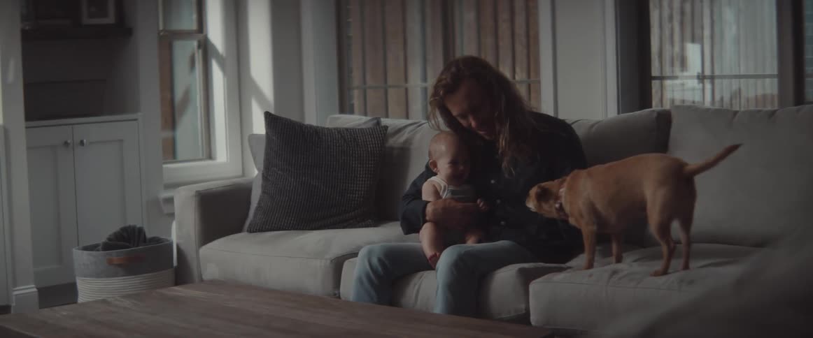 A dad bouncing his baby on his leg pets the dog when it jumps up onto the couch