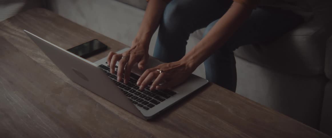 A person sits on a couch and leans to type on the laptop on the table in front of them