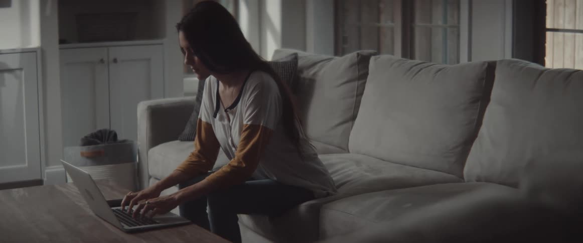 A woman sits on a sofa in a living room leaning forward to type on her laptop
