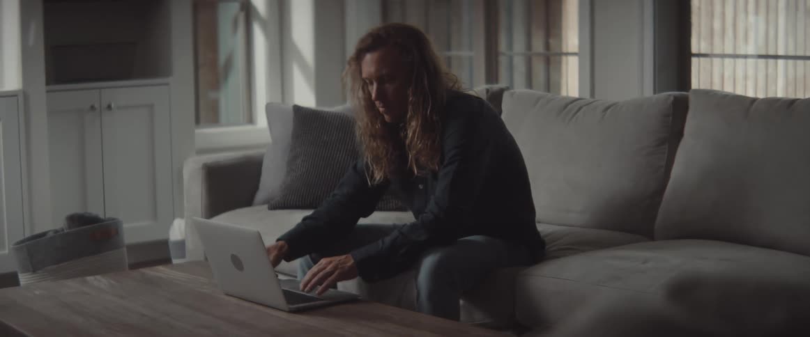 Man texts on mobile phone then types on laptop as he sits on couch in living room