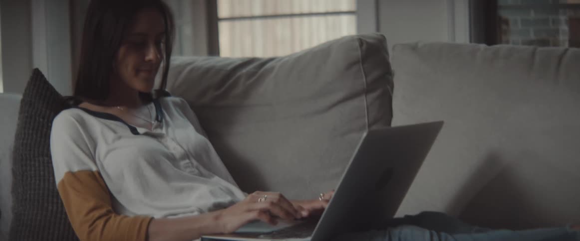 Woman with dark hair white sweatshirt and jeans sits on grey couch and types on laptop