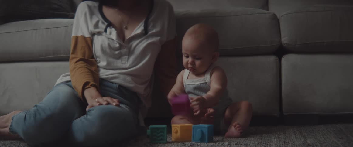 A baby plays with toy blocks as a woman sits besides them on the floor in the living room of a house