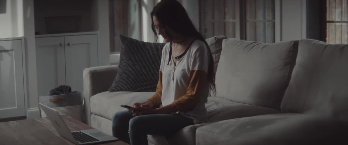 A young woman glances at the screen of a laptop before typing on a smartphone while sitting on a sofa in the living room of a house during the day