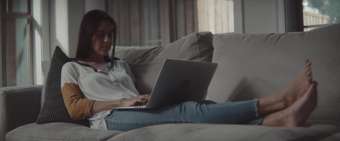 A woman sitting on a couch typing on a laptop with a wall and window in the background