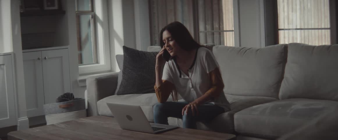 Woman with long dark hair and sweatshirt sitting on couch talks on smartphone and uses laptop in living room at home