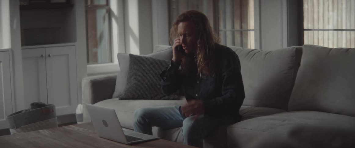 A man at home sits on a couch in front of his laptop and talks on his cell phone