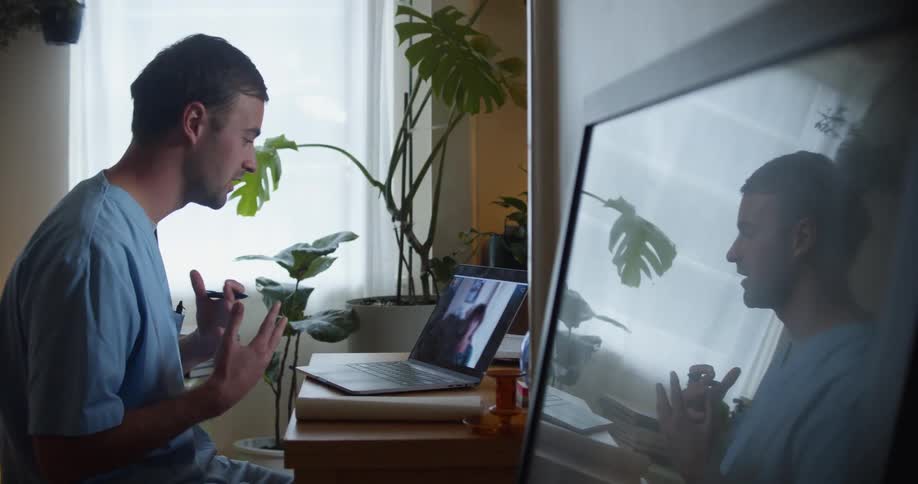 Man sits in home office talking with another via video call on laptop and taking notes