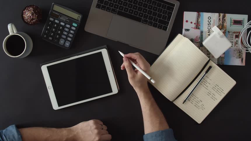 A man sits at a desk with a table computer and cup of coffee and writes in a book