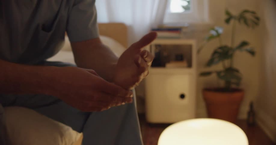 A person sits on a bed and rubs their hands together