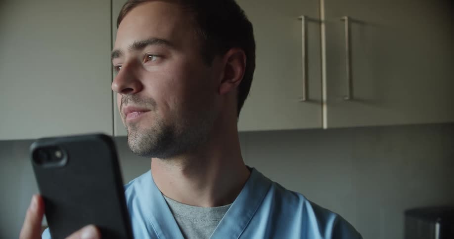A man with scrubs in a room holding up a smart phone with a wall and cupboards in the background