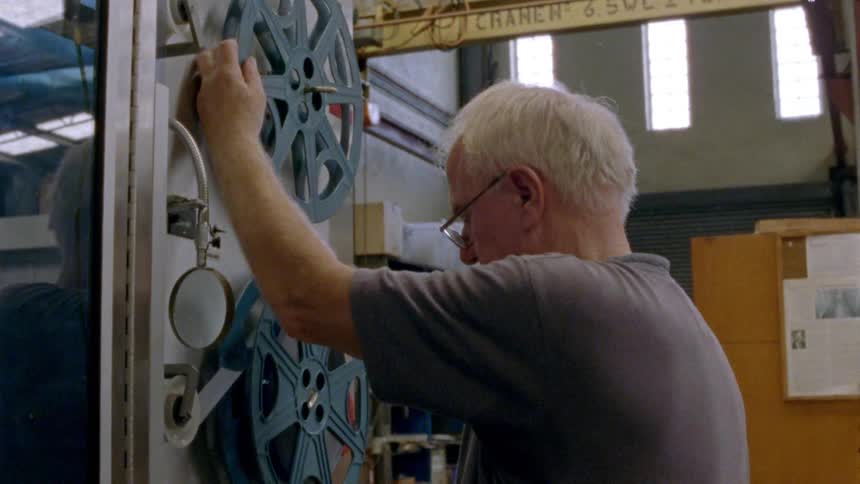 A man with glasses in a room putting a film reel on a machine