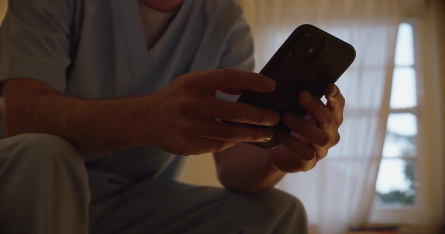 Man with light blue scrub tunic sits down and uses iphone smartphone