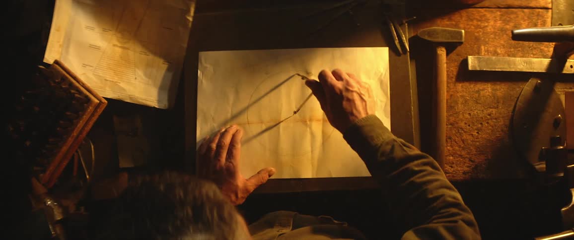 A man uses a compass to draw a circle on a sheet of paper in a dimly lit workshop