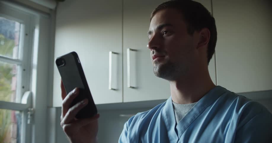 A nurse holds a smartphone in front of his face while speaking and gazing out of a nearby window