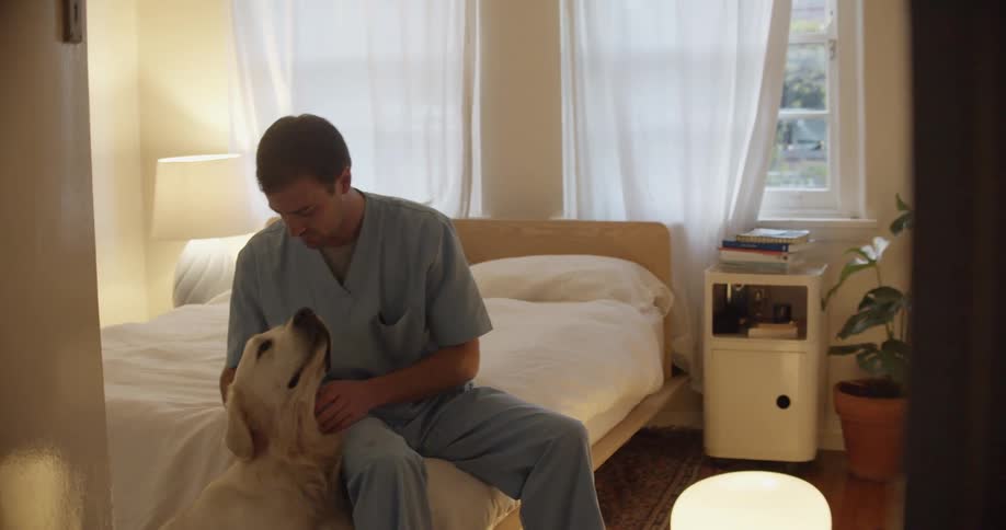 A man in nursing scrubs sits on the end of his bed and hugs and kisses his large white dog