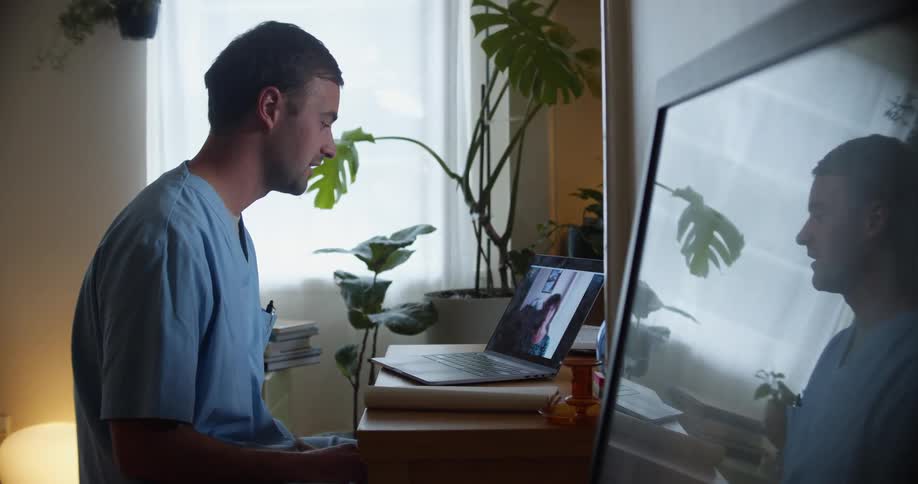 A man sitting at a desk looking at a laptop with a wall and window in the background
