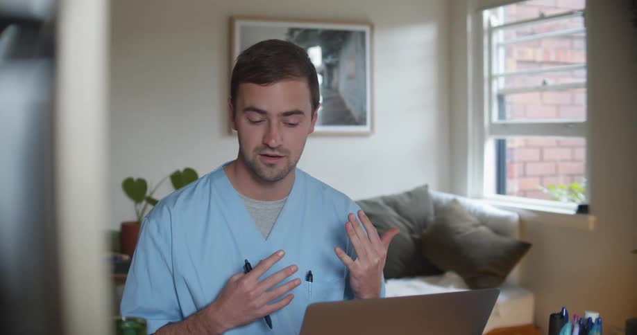 A man in a room wearing scrubs talking on a laptop with a wall and window in the background