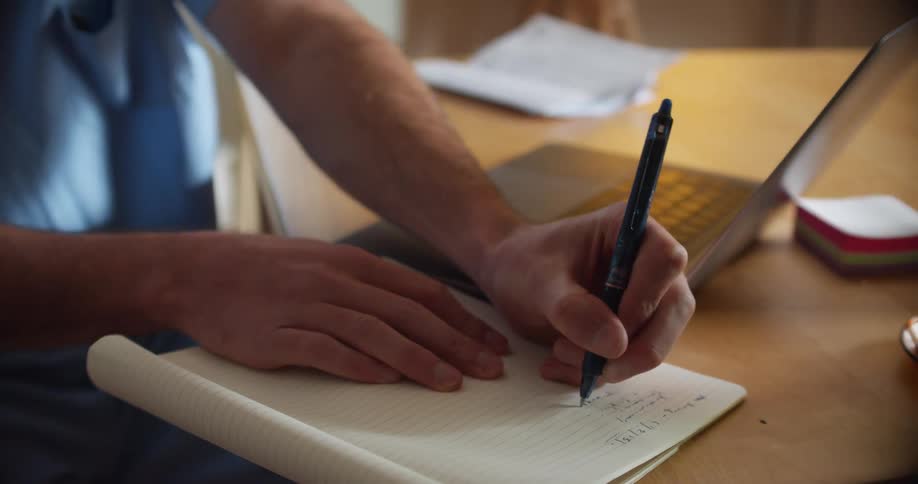 A person sits at a table writing in a notebook with a laptop beside them