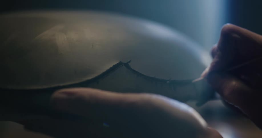 Person uses pointed wooden skewer to sketch line onto clay in dim room light by beam of light