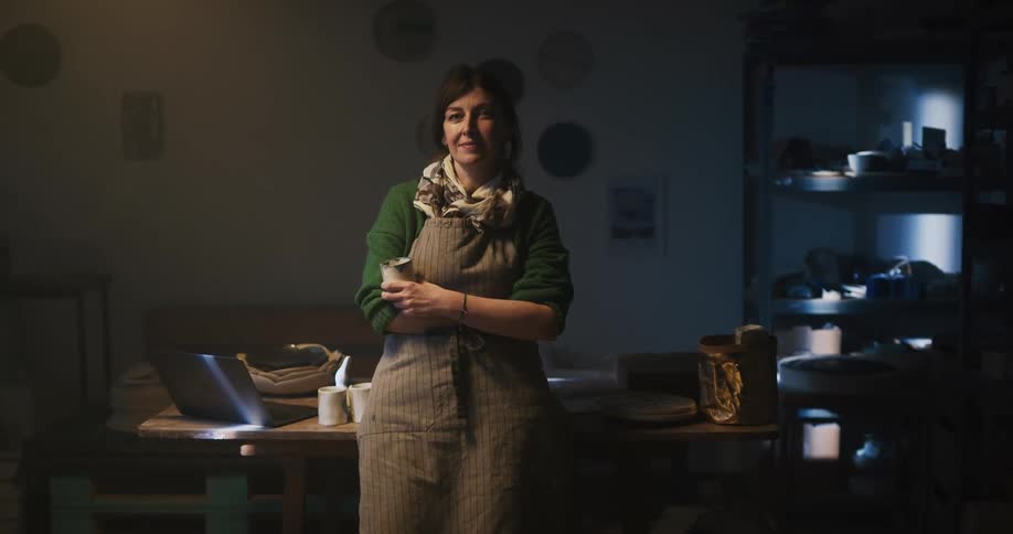 A woman leans against her desk and holds a mug and smiles