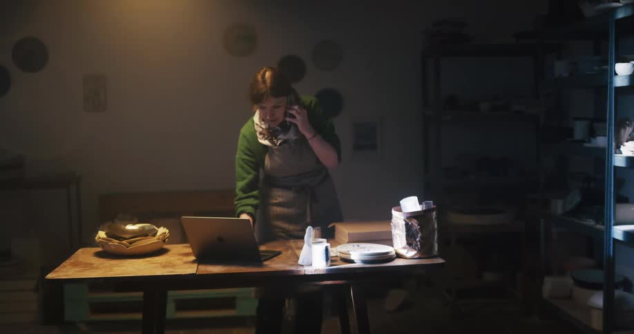 A woman walks around her desk while talking on the phone then stops to check her laptop