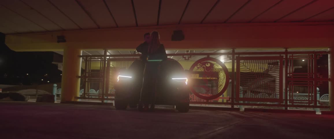 A man and a woman hugging in front of a vehicle with headlights