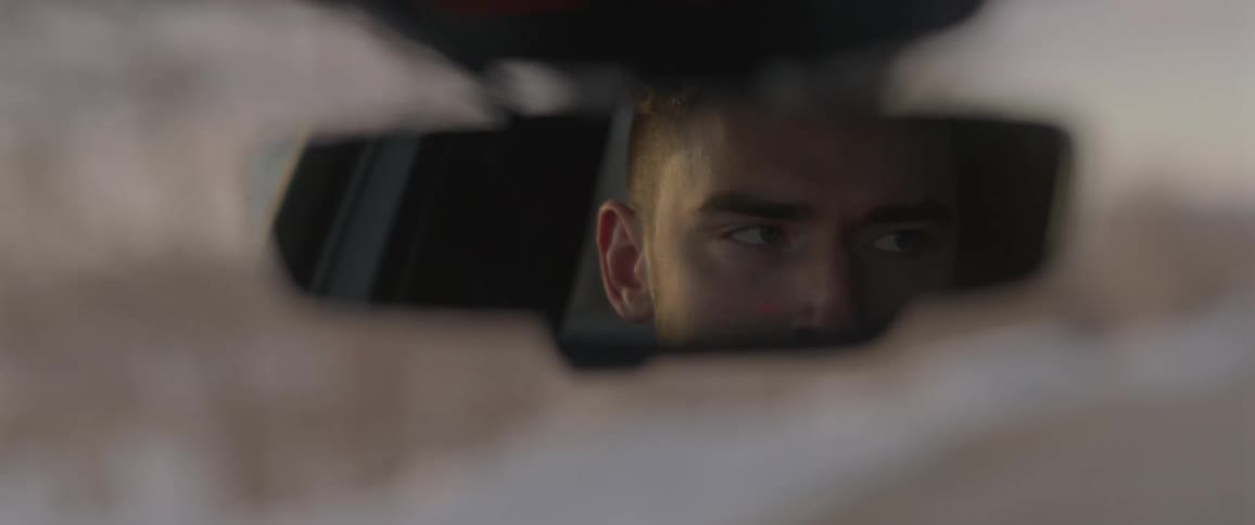 A man in a vehicle with his reflection in the rearview mirror with the sky and clouds in the background
